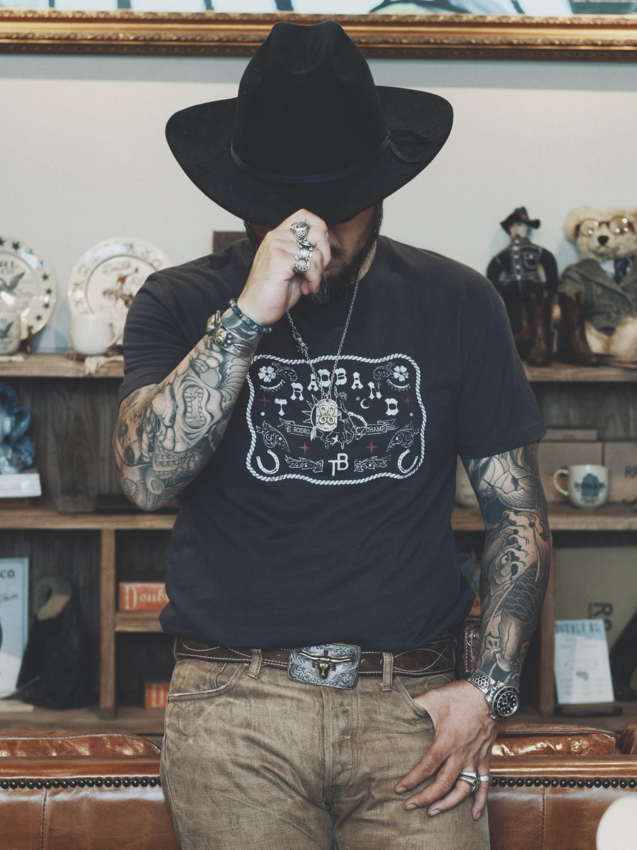 Rugged-looking man with cool Japanese style tattoos wearing a black cotton Distressed Vintage Cowboy Belt Buckle Graphic T-Shirt, vintage RRL pants, and leather belt with belt buckle picturing bull's head