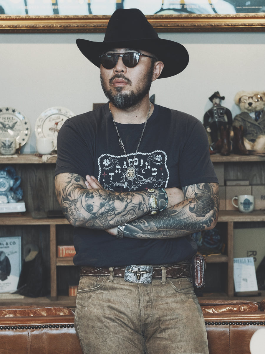 Rugged-looking man with cool Japanese style tattoos wearing a black cotton Distressed Vintage Cowboy Belt Buckle Graphic T-Shirt, vintage RRL pants, and leather belt with belt buckle picturing bull's head